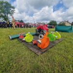 500562422_728984039477120_1315881667073960017_n Strażacy stoją na trawie obok czerwonych wozów strażackich. Na pierwszym planie kolorowe węże strażackie na drewnianej platformie. W tle widoczne zielone drzewa i niebo z białymi chmurami.