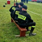 500641358_728989439476580_6645669675321909816_n Trzech strażaków w czarnych mundurach siedzi tyłem na ławce. Na plecach mają żółte napisy STRAŻ. Tło to zielona trawa i sprzęt strażacki.