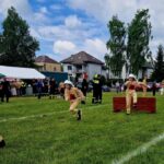 500657654_728981106144080_6659595351845741602_n Młodzi strażacy w beżowych strojach i kaskach biegną po trawie. Tło to drzewa i namioty, pogoda pochmurna. Obok skaczą przez przeszkody.