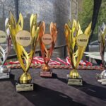 500767679_727959016246289_5258109489684021585_n Na czarnym stole stoją trofea: złote, srebrne i brązowe, oznaczone jako 1, 2, 3 miejsce. Z tyłu widać medale. Tło jest lekko rozmyte, z fragmentem zieleni.