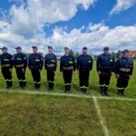 500841149_728990029476521_3928600294227666089_n Grupa strażaków w czarnych mundurach stoi na zielonej trawie. Niebo jest jasne z białymi chmurami. W tle widać dom i pojazdy.