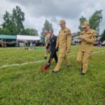 500841537_728973802811477_6165147004610743444_n Grupa osób w beżowych mundurach chodzi po trawie z narzędziem pomiarowym. W tle namiot i drzewa. Szeroka, zielona przestrzeń.