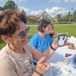 500905043_728062679569256_4969317950979757829_n Osoba trzyma ozdobiony kamień z białym kotem, tło to zielona trawa i domy. Na stole widać przybory do malowania.