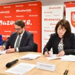 Trzy osoby podpisują dokumenty w sali z czerwonym banerem Mazowsze. Kobieta w niebieskiej marynarce, mężczyzna w garniturze i kobieta w czarnej marynarce siedzą przy drewnianym stole. Obok stoją mikrofony i tabliczka z napisem