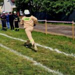 500974330_728976396144551_4460479684340759374_n Osoba w stroju strażackim biegnie po trawie. Na lewo kilku strażaków w czarnych mundurach. W tle drzewa i ogrodzenie.