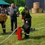 501031683_728979796144211_635280367103430616_n Osoba w strażackim stroju wylewa wodę z wiadra do hydrantu na trawie. Obok obserwują inni strażacy w beżowych koszulkach. W tle namiot i bele siana.