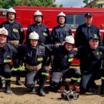 501040988_728978329477691_560094425884232873_n Grupa strażaków w czarnych mundurach stoi przed czerwonym wozem strażackim. W tle zielone drzewa. Obok stoi kobieta i mężczyzna w garniturze.