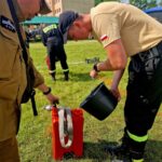 501050921_728980886144102_5440681107232356352_n Dwóch mężczyzn w uniformach strażackich nalewa wodę z czarnego wiadra do czerwonego pojemnika. Tło to trawiaste pole z niskim budynkiem i drzewami. Słońce oświetla scenę, dając jasne, żywe kolory.