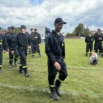 501052511_728983659477158_5494547496674565880_n Grupa strażaków w czarnych mundurach z żółtymi elementami stoi na trawie. Jeden chłopiec siedzi na ziemi. W tle niebo z chmurami i kilka budynków.