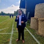 501055052_728976806144510_707138225993811224_n Mężczyzna w garniturze z mikrofonem stoi na trawie. Obok niego trzy bele siana. W tle niebo z chmurami i ludzie.