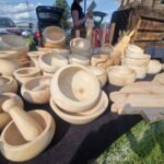 501066055_728062819569242_5310673632065364333_n Na zdjęciu widać drewniane naczynia i moździerze na stoisku. Kolory są naturalne, beżowe, w tle samochody. Tło zielone, trawa i niebo.