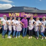 501071691_728062399569284_2264010163252690854_n Grupa ludzi w białych koszulkach i fioletowych fartuchach stoi przed namiotem z napisem KGW Porębianki. Tło to zielona trawa i niebo z chmurami. Osoby mają na głowach kwiatowe wianki.