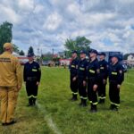 501072039_728991276143063_2090670034441808534_n Strażacy w mundurach ćwiczą na zielonym polu. Niebieskie niebo z chmurami w tle. Wokół zielone drzewa i budynki.