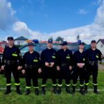 501090743_728978099477714_9075476735899346931_n Grupa strażaków w ciemnych uniformach stoi na trawie. W tle dwa czerwone wozy strażackie i domy pod błękitnym niebem z chmurami.