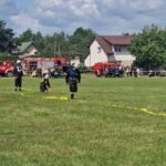 501168789_728983389477185_9205155513228192123_n Strażacy ćwiczą na zielonej łące z żółtymi wężami strażackimi. W tle czerwone wozy strażackie i białe domy. Na niebie białe chmury.