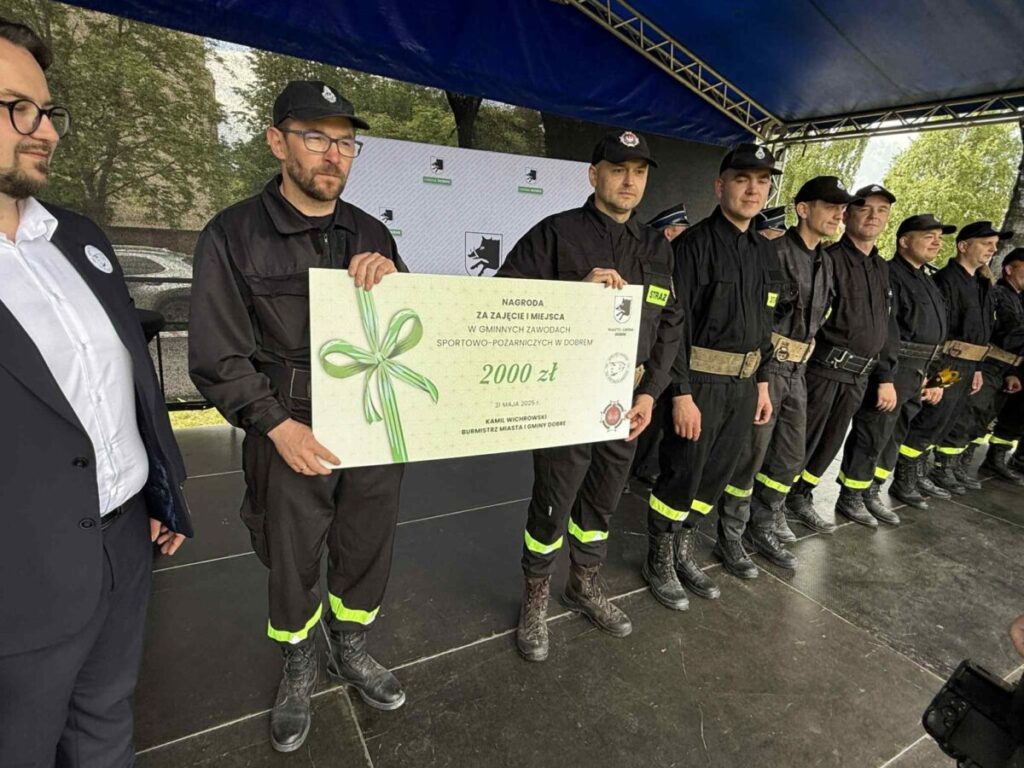 Grupa osób w czarnych mundurach stoi na scenie pod niebieskim namiotem. Jedna osoba trzyma duży biały czek. W tle drzewa i logo organizatora.