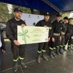 501172755_728972579478266_8097564159870550498_n Grupa osób w czarnych mundurach stoi na scenie pod niebieskim namiotem. Jedna osoba trzyma duży biały czek. W tle drzewa i logo organizatora.