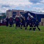 501193197_728984832810374_7771450152903910445_n Strażacy w strojach ochronnych przygotowują się do zawodów na trawie. W tle widać namiot, budynki i chmury na niebie. Po prawej stronie są ustawione bele siana.