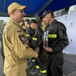 502433116_728982642810593_4333541637283142369_n Strażacy w czarnych mundurach i żółtych naszywkach STRAŻ otrzymują gratulacje. Tło to niebieski namiot i biała ścianka z logo gminy. Atmosfera jest uroczysta.