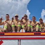 502465559_728970826145108_4252981547171172790_n Pięciu młodych strażaków siedzi na dachu pojazdu. Noszą beżowe mundury z żółtymi paskami, prezentując gesty rękami. Tło z niebieskim niebem i białymi chmurami.