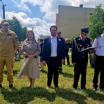 502467273_728971322811725_7805352531094410639_n Grupa pięciu osób stoi na tle zielonej trawy. Trzy osoby w mundurach, jedna w eleganckiej sukience. Tło tworzą drzewa, niebo i budynek.