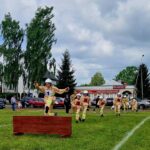 502486008_728969189478605_6113056107105635344_n Strażak w żółtym mundurze skacze przez przeszkodę na trawie. W tle kilku strażaków, budynki i drzewa. Niebo zachmurzone.