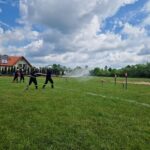 502494434_728970486145142_3175262799664536599_n Grupa strażaków w czarnych mundurach ćwiczy na zielonej łące. Tło to dom z czerwoną dachówką i chmury na niebie. Instruktor w beżowym stroju stoi z boku.