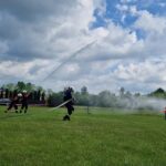 502561932_728970579478466_6664790466776980529_n Strażacy w hełmach ćwiczą z wężami na zielonym polu. W tle niebieskie niebo z chmurami. Z boku stoi dom i drzewa.