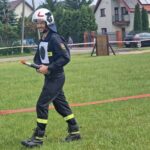 502581448_728970686145122_5391135640381068501_n Mężczyzna w stroju strażaka z uśmiechem trzyma narzędzie na trawie. W tle domy i zaparkowane auta. Dzień, zielona trawa.