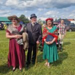 502584498_728061736236017_766430491128585460_n Trzy osoby w stylowych strojach na tle zielonej trawy i nieba z chmurami. Kobieta po lewej w bordowej sukni i kapeluszu, mężczyzna w garniturze z melonikiem, kobieta po prawej w zielonej sukni z czerwonym kwiatowym