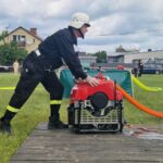 502612321_728985306143660_2306889197129021338_n Na zdjęciu strażak w czarnym mundurze obsługuje czerwoną pompę wodną na trawie. W tle widoczne są domy i samochody. Niebo jest pochmurne.