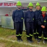 502654700_728973529478171_9151379614738704403_n Czwórka osób w czarnych mundurach i zielonych hełmach pozuje na tle białego namiotu. Tło zawiera napis OSP MLECIN. Trawa pod stopami jest zielona i gęsta.