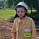 502732020_728975039478020_9062574027479626218_n Na zdjęciu młoda osoba w beżowym stroju strażaka z białym kaskiem. Tło to zielona trawa i drzewa. Na kombinezonie widoczny jest napis STRAŻ.