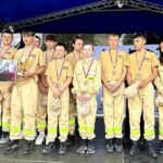 502766332_728966956145495_7490203822074545518_n Grupa młodzieży w mundurach strażackich stoi na scenie, trzymając medale. Obok dorosły w garniturze. Tło w kolorze niebieskim.