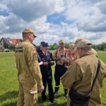 503110937_728970632811794_140251968807057725_n Grupa strażaków na zielonym polu, ubrani w jasne mundury pod pochmurnym niebem. W tle domy i drzewa. Trwa narada.