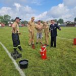 503215273_728974416144749_4979983123250418462_n Grupa osób w beżowych i czarnych ubraniach stoi na zielonym boisku. Na trawie widać czerwony sprzęt strażacki. W tle są wozy strażackie i niebo z chmurami.