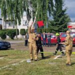 503244814_728975349477989_3268825299840778948_n Na zielonej trawie stoją strażacy w beżowych mundurach z czerwonymi flagami. W tle biały budynek i czerwone auta strażackie. Dzień jest słoneczny.