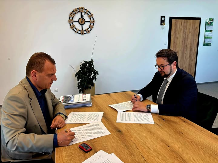 Dwóch mężczyzn siedzi przy drewnianym stole i podpisuje dokumenty. Ściana jest biała, z zegarem i rośliną w tle. Jeden mężczyzna ma garnitur, drugi jasną marynarkę.