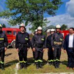 503445244_728977986144392_1313545090111157810_n Grupa strażaków w mundurach stoi na trawie z czerwonymi wozami strażackimi w tle. Obok nich, mężczyzna w garniturze. Na niebie widać chmury i drzewa.