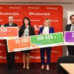 Pięć osób trzyma kolorowe tablice z kwotami dla Gminy Dobre na tle czerwonego baneru z napisem Mazowsze. Ubrani formalnie, stoją w szeregu. Tablice są czerwone, zielone i fioletowe.