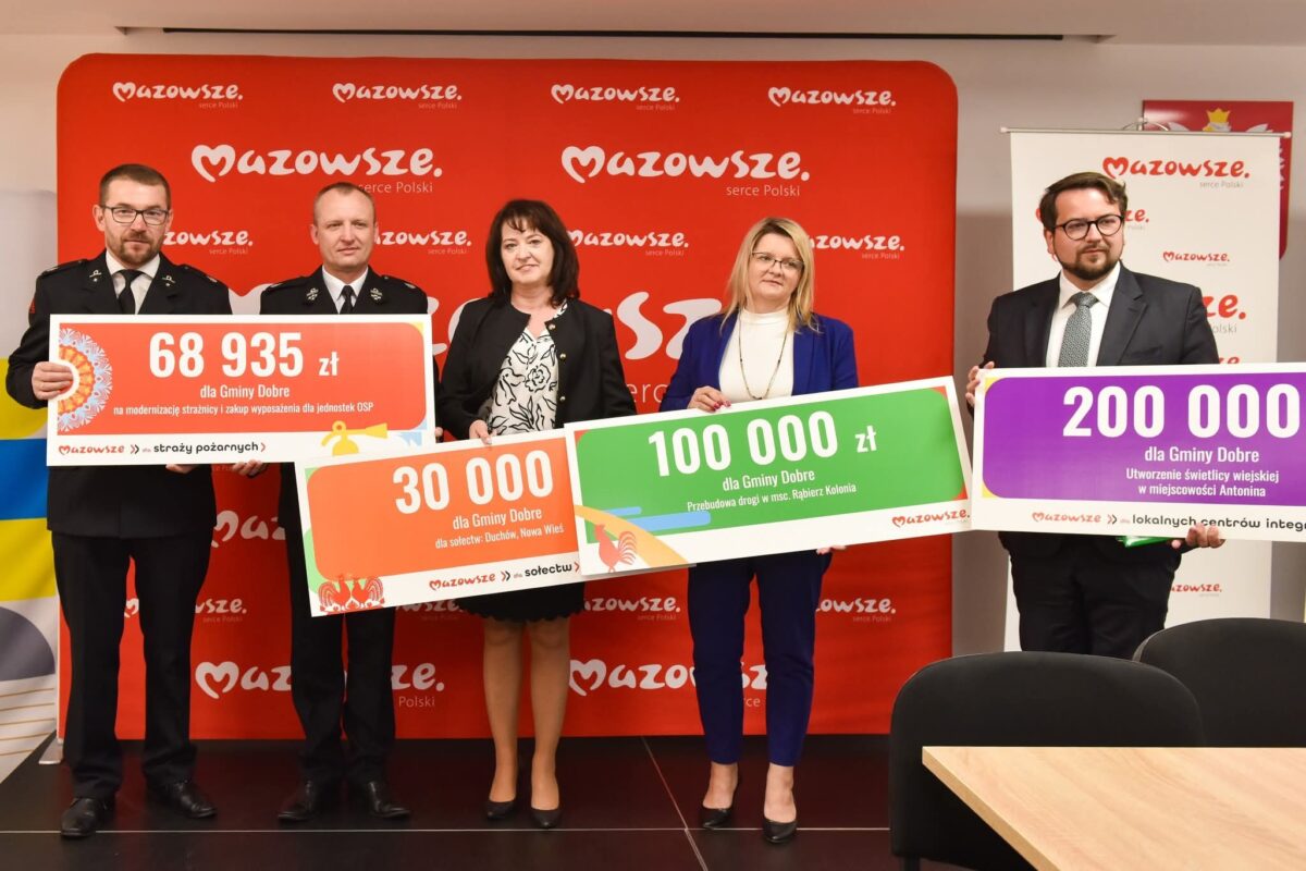 Pięć osób trzyma kolorowe tablice z kwotami dla Gminy Dobre na tle czerwonego baneru z napisem Mazowsze. Ubrani formalnie, stoją w szeregu. Tablice są czerwone, zielone i fioletowe.