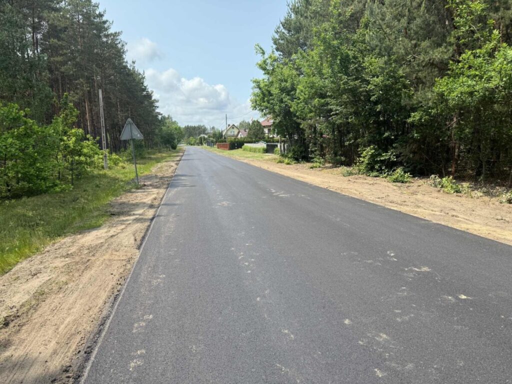 Asfaltowa droga przez las, po lewej znaki i drzewa, po prawej domy. Jasne, letnie niebo z chmurami. Tło zielone.
