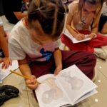 Na zdjęciu kilka dzieci rysuje na kratkowanych arkuszach, siedząc na jasnej podłodze. Każde dziecko trzyma ołówek. Atmosfera skupiona, tło ciemne.