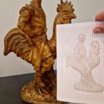 Brązowa rzeźba przedstawiająca mężczyznę na kogucie. Tło jest jasne, a obok znajduje się rysunek konturowy na kartce. Kolory to głównie brązy i biel.