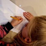 Dziewczynka w różowej bluzie rysuje ołówkiem na kartce w kratkę. Włosy jasne, tło jasne. Zbliżenie na dłonie.