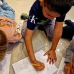 Chłopiec rysuje na kartce w kratkę leżąc na podłodze. Ma na sobie ciemną koszulkę i szare spodnie. Tło jest jasne, podłoga beżowa.