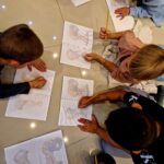 Dzieci rysują na podłodze, trzymając ołówki nad zeszytami z kolorowankami. Tło jest jasne i jednolite. Widoczne są kolorowe ubrania dzieci i skupiona atmosfera.