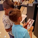 Dwóch chłopców ogląda rzeźby w galerii sztuki, trzymając notatki. Mają ubrane kolorowe koszulki. W tle są różnorodne rzeźby na szarych postumentach i jasna podłoga.