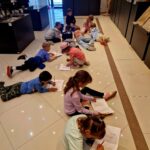 Grupa dzieci leży na jasnej podłodze i pisze w muzeum. W tle widać rzeźby na ciemnych postumentach. Ściana jest jasna, co dodaje przestrzeni.