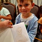 Chłopiec w jasnoniebieskiej bluzie siedzi na krześle. Trzyma kartkę z konturem postaci na siatce. W tle są inne osoby i obrazy.